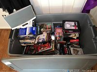 Large gray plastic bin filled with assorted DVDs and VHS tapes, Bose speaker placed next to the bin.