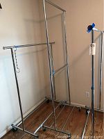 Two large metal clothing racks and a smaller one positioned on a wooden floor near a wall and window in a room.