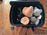 Photo showing inside of black plastic storage bin with two round embroidered woven baskets, pair of vintage blackboxing gloves, and one handmade grey bear doll with black bow tie