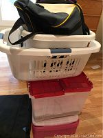 Stacked shot showing one white plastic laundry basket on top of two clear plastic storage bins, with black and yellow duffel bag visible in the basket