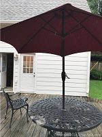 Photo of the round black iron lattice patio table with maroon umbrella fully open and one black metal chair on wooden deck.