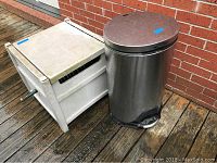 Photo of a white hard plastic garden hose reel box with garden hose and a round stainless steel trash bin with pedal, placed outside on a wooden deck against a brick wall.