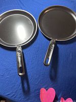 Top view of two non-stick crepe/pancake pans on blue fabric background, showing smooth cooking surfaces and black handles with ventilation holes.