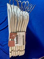 Photo of 8 new white wooden hangers with metal hooks held together by a branded paper band stating 'Solutions - Hotei Hanger - White'.