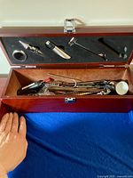 Wooden hinged bar tool box open showing black foam insert in lid holding four bar tools: two metal pour spouts, muddler, and metal stirrer, with additional bar tools in lower compartment.