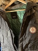 Detailed photo showing the exterior of the brown Bench parka with its zippered front and faux fur hood trim, along with an emblem patch on the sleeve.