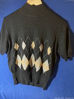 Black short-sleeve sweater with cream and yellow argyle pattern on the front, displayed on hanger against blue background.