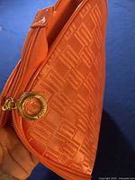 Close-up of orange cosmetic bag showing embossed logo pattern and gold-tone zipper pull with Estee Lauder engraving.