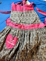 Full apron laid flat showing pink upper area, patterned skirt section, pink waistband, and heart-shaped pink pocket embroidered with Kitchen Diva logo.