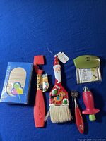Top view of lot showing all five items arranged in line on a blue background: wooden soap holder box, red bottle opener, Santa Claus brush, metal tool with red handle, boxed knife sharpener
