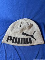 Gray Puma beanie with large black Puma text and puma cat logo on front laid flat on blue background.