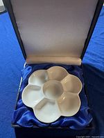 Front view of a white ceramic paint palette shaped like a flower with seven wells placed in a cushioned display box with blue satin lining.