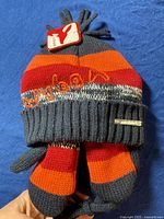 Photo of child's knit hat and mittens set on blue fabric background, showing striped red, orange, maroon, and gray colors and Aardvark brand tag.
