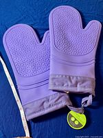 Pair of purple silicone oven mitts laid flat on a dark blue fabric showing the textured silicone surface and fabric cuffs with hanging loops. A tape measure indicates mitt length approximately 11 inches.