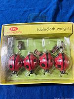 Front view of the packaged ladybug lane tablecloth weights showing four ladybug shaped weights with clips inside yellow packaging.