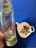 Joie tea bag squeezer in original packaging alongside porcelain tea bag plate with a red flower design on a blue cloth background.