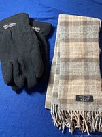 Black fleece Thinsulate gloves and beige-brown plaid Merino wool scarf laid on blue background.