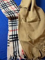 Photo showing two men's scarves: one beige with fringed ends and one plaid with beige, black, white, and red colors.