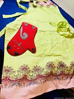 Yellow apron with a decorative pink and brown border on the bottom, laid flat showing neck strap and waist ties, with red cat-shaped oven mitt placed on top.