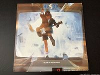 Front album cover of AC/DC Blow Up Your Video vinyl record showing the band member with guitar in schoolboy outfit among broken glass effect.