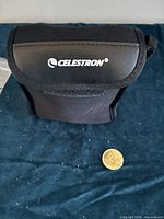 Black soft carrying case with Celestron logo, photographed with small coin for size reference on fabric background.