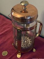 Medium sized Bodum coffee press with copper lid, brass handle and feet, glass body, and metal plunger.