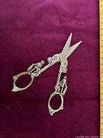 Top view of the silver-plated herb scissors showing detailed ornamental figures on handles and etched floral designs on the blade area with a measuring tape showing length about 15 cm.