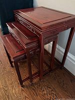 View of three red wood nesting tables stacked, showing the largest table on top with brass circular inlay and carved geometric designs on apron sides.
