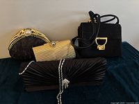Group view of 4 bags: Black Coccinelle suede purse, black ruched satin baguette evening bag with rhinestone chain, two small gold-tone frame clutches, one with pearl accent.