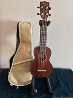 Full view of Gretsch ukulele on a black stand next to beige Gretsch gig bag, showing the body and fretboard.