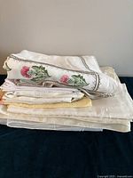 Stack of folded tablecloths and napkins showing various embroidered and plain linens in white and cream shades with floral detail.