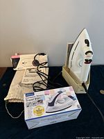 Lot shows Braun steam iron positioned on a cream-colored stand, alongside a compact white and pink Sanyo iron with cord and instruction manual, and a boxed Oxo compact steam/dry travel iron providing visual details of condition and packaging.