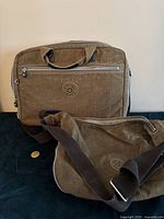 Photo showing front view of khaki Kipling briefcase/computer bag with zipped front pocket and Kipling logo. Crossbody purse with strap partially visible.