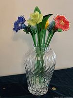 Full view of large clear crystal vase holding colorful glass flowers showing variety of blue, yellow, and red flowers with green stems.