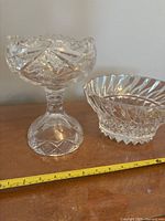 Photo showing two heavy leaded crystal bowls side by side. One is a tall pedestal bowl with a scalloped edge and intricate cut glass design; the other is a shorter bowl with vertical and starburst cut patterns on the sides and base. A measuring tape is included showing approximate size.