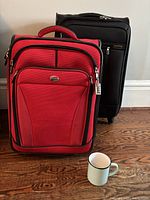 Two upright suitcases, a red American Tourister and a black suitcase, shown standing on wooden floor next to a coffee mug.