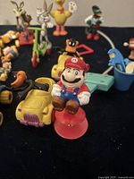 Close-up of assorted McDonald's character toys featuring Super Mario figurine in front with various Looney Tunes and other characters behind on a black surface.