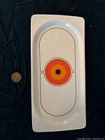 Top view showing small rectangular white ceramic tray with rounded oval inset and orange-red-black concentric circle design in middle, coin placed for scale.
