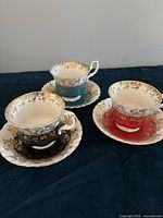 Three Royal Albert bone china teacups with matching saucers in black, teal, and red, displayed on a dark surface.
