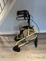 Side view of yellow 3-wheeled shopping walker with plaid canvas bag, black wheels, and hand brakes on handles.