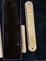 Silver and brass mezzuzah case outside of its box with prayer scroll beside it on a dark fabric surface.