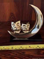 Front view of two small brass owls sitting on a crescent brass base attached to a black rectangular stand with measuring tape showing size approximately 4 inches wide behind the item.