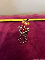 Small Murano glass clown baby figurine seated in sterling silver high chair, showing detailed colors and structure against a maroon backdrop with a measuring tape in the background.
