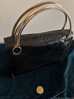 Front angled view of a black snakeskin leather handbag with metallic triple gold-tone handles, placed on a dark surface next to a coin for size reference.