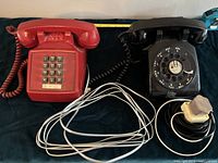 Two vintage telephones side by side, one black rotary dial and one red push-button, showing cords and adaptor.