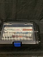 Front view of the Dremel accessory case showing the upper tier with multiple grinding stones, sanding drums, and bits arranged in a white plastic holder behind a clear hinged cover.