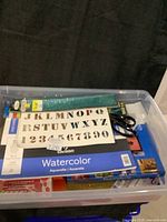 Top view of clear plastic container showing Canson watercolor paper pad, alphabet stencil sheet with letters and numbers, scissors, and other craft supplies inside.