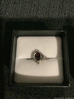 Front view of the silver ring in black and white cushioned box showing the marquise-shaped dark red stone.
