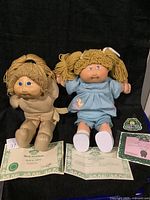 Two Cabbage Patch Kids dolls side by side with birth certificates and green Cabbage Patch Kids tags laid out in front.