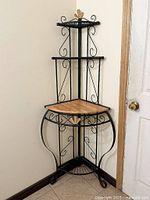 Overall view of the 4 tier corner bakers rack with wood-finish middle shelf and black metal frame with decorative scrollwork and gold fleur-de-lis accents.